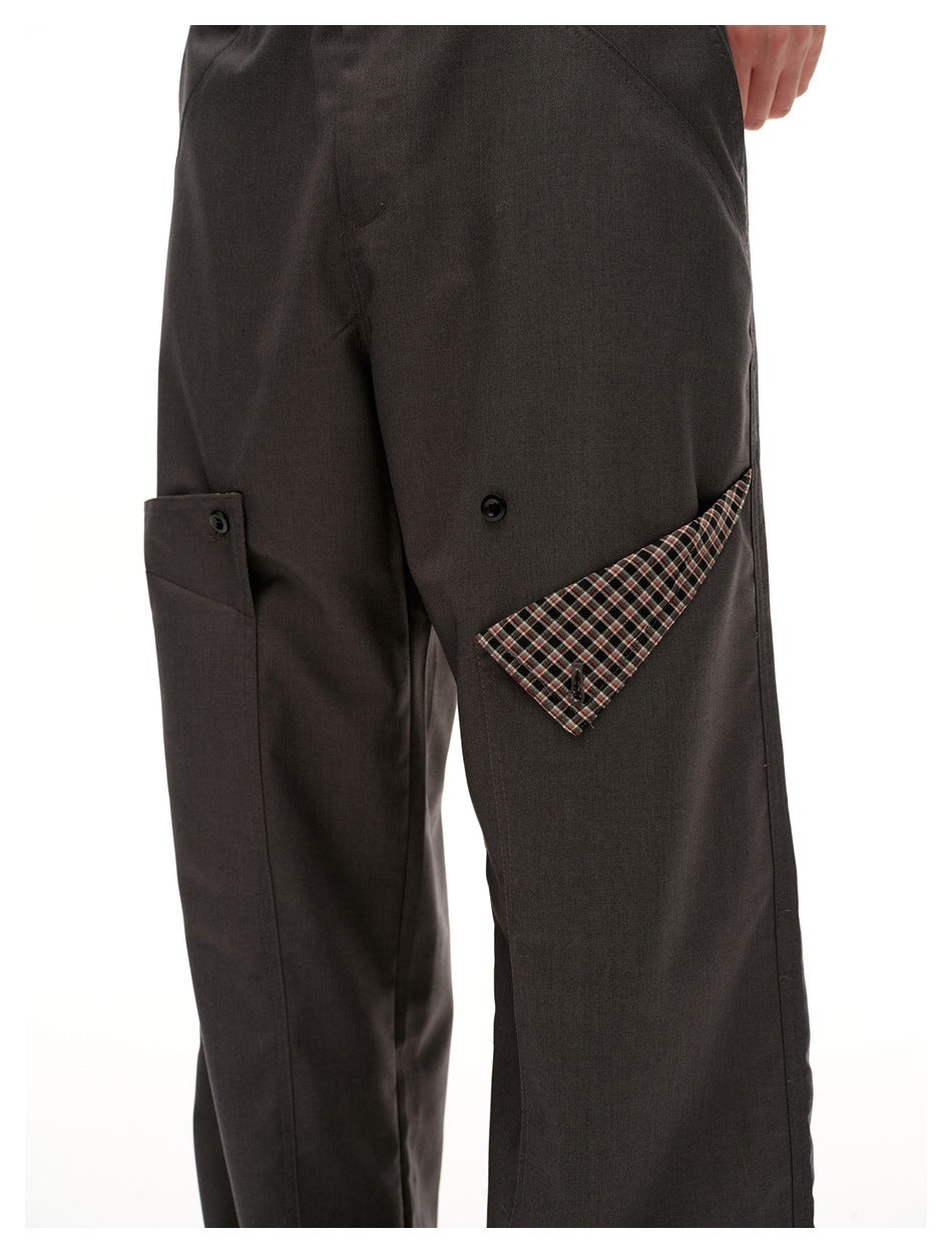 Close-up of charcoal trousers showing a buttoned utility pocket and a triangular plaid fabric accent.