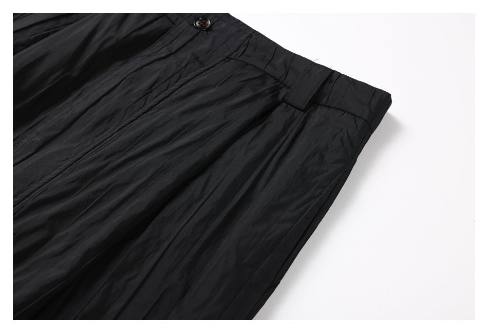 Close-up of black pleated trousers showing the waistband, button, belt loop and crinkled fabric.