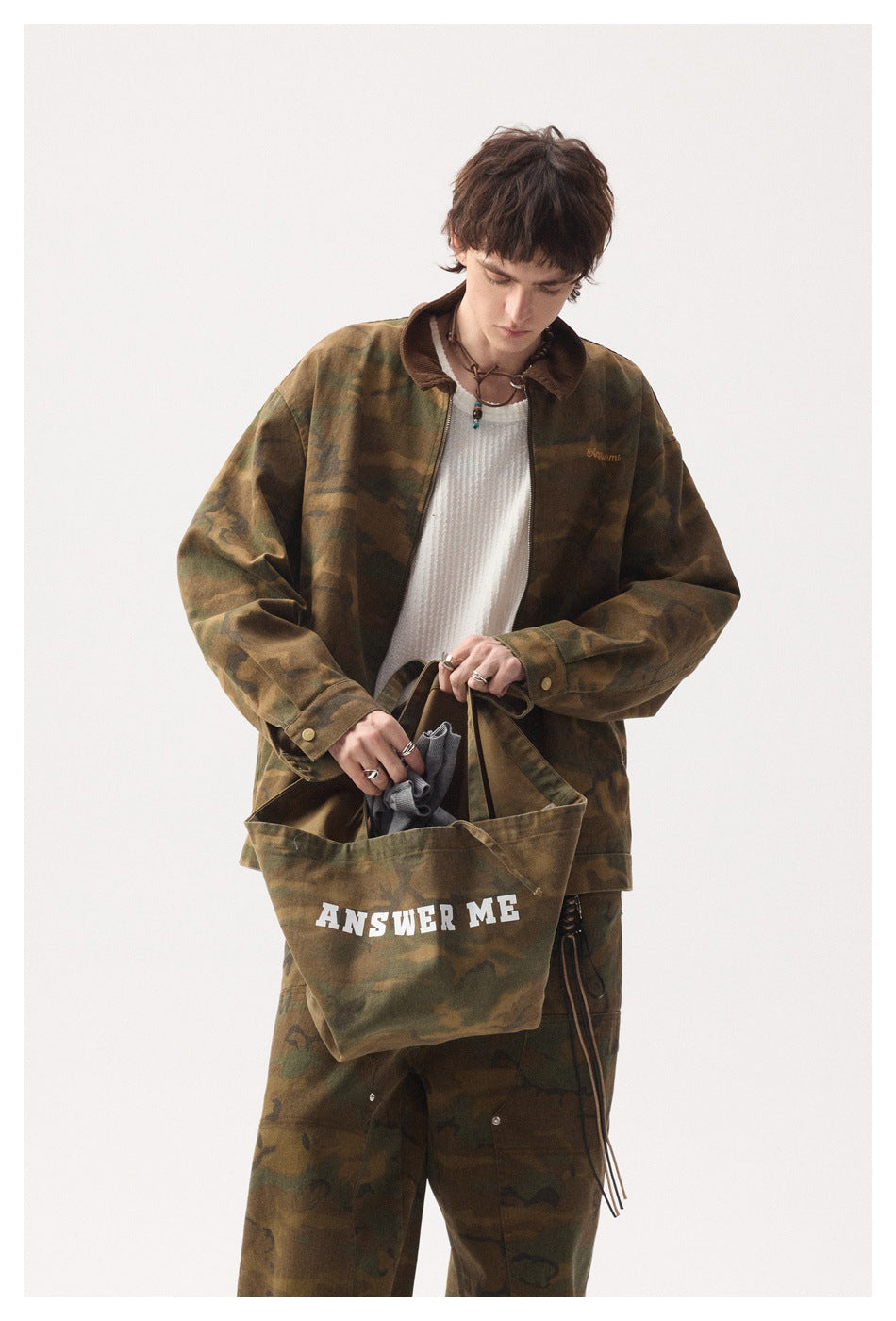 Model wearing camouflage jacket with brown collar while holding a camouflage tote bag printed ANSWER ME
