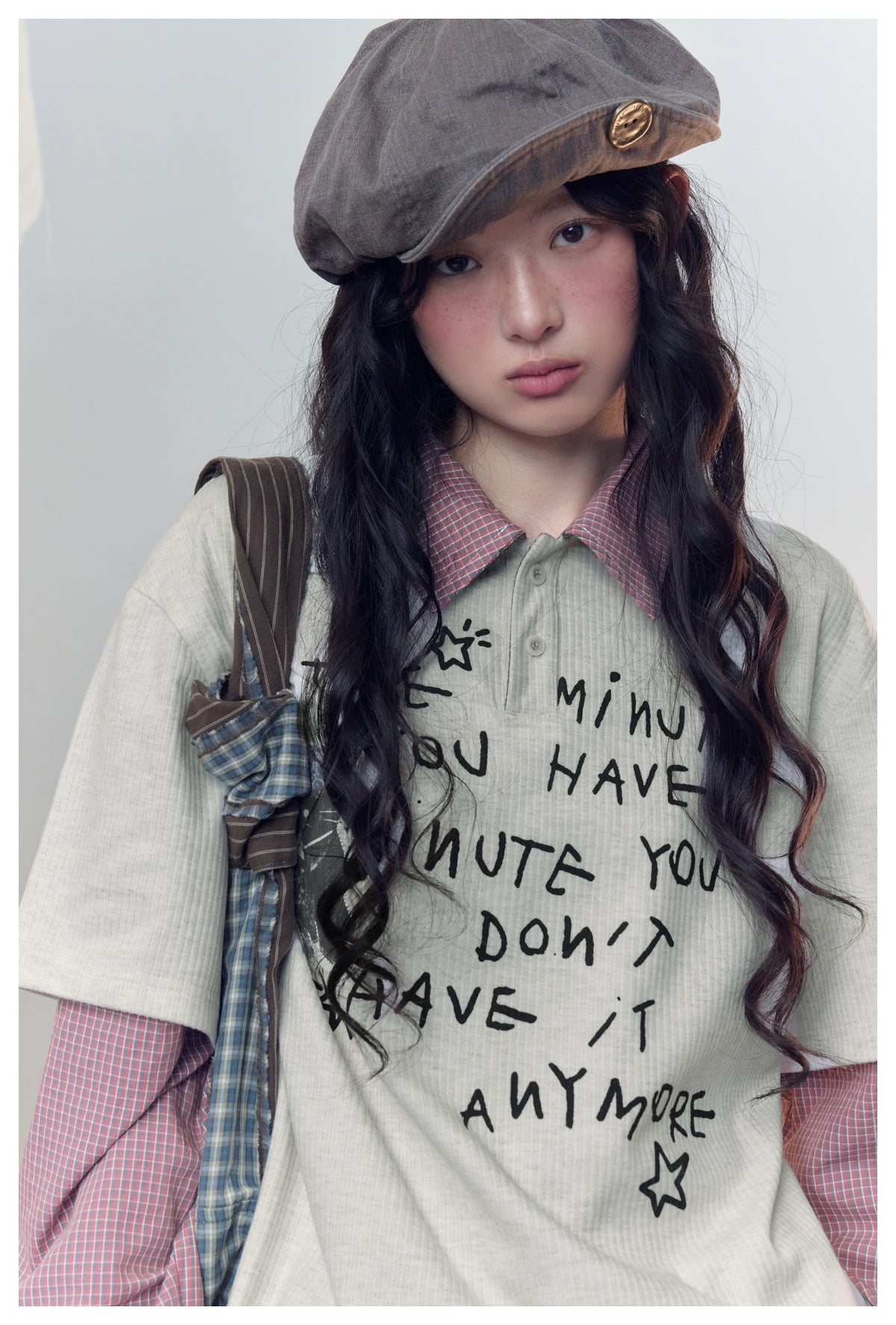 Close-up of a model in a pale gray graphic tee over a pink checked shirt, with a gray-brown buttoned cap.