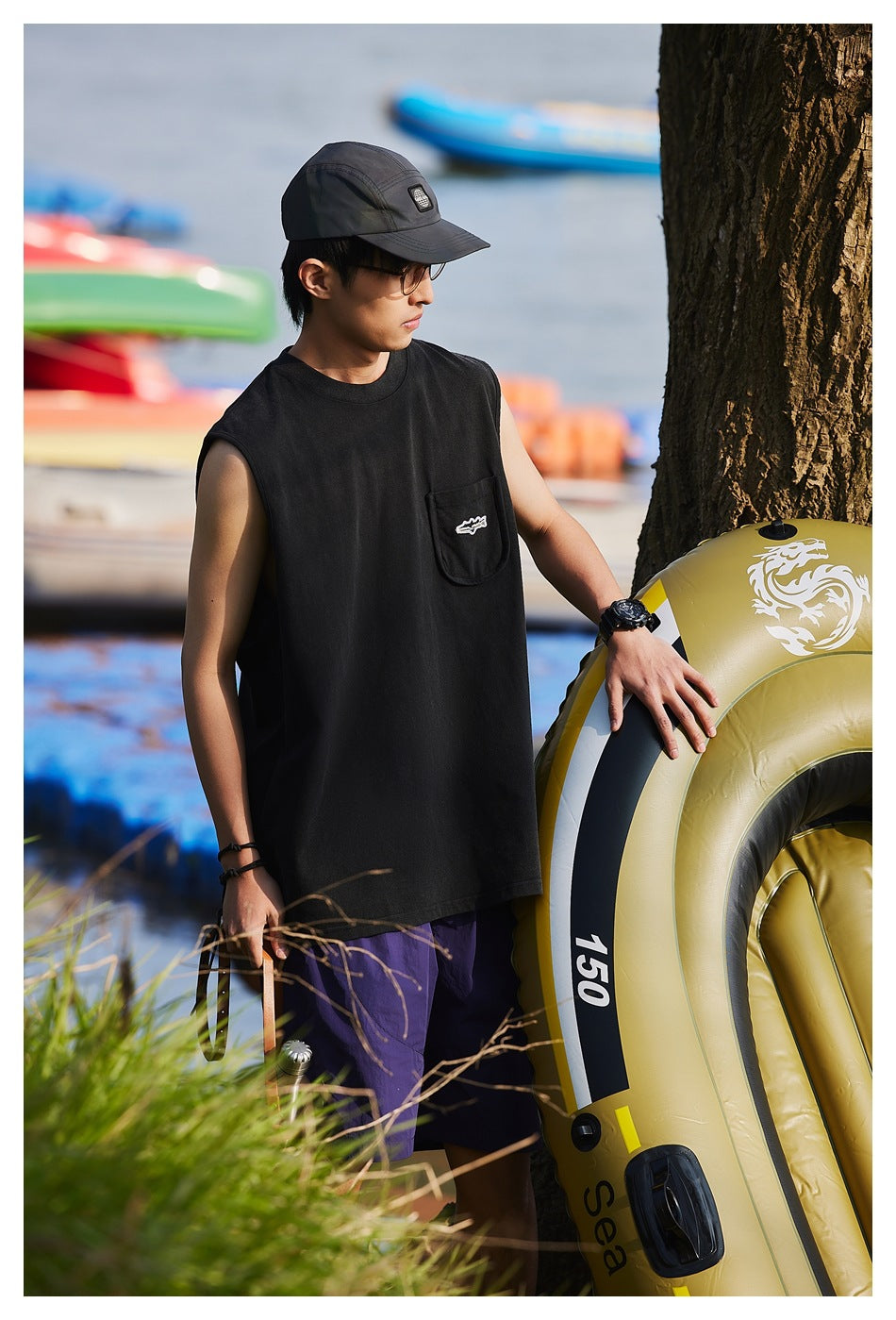 Side view of a model in a black sleeveless pocket tank top and purple shorts by a lake with an inflatable boat
