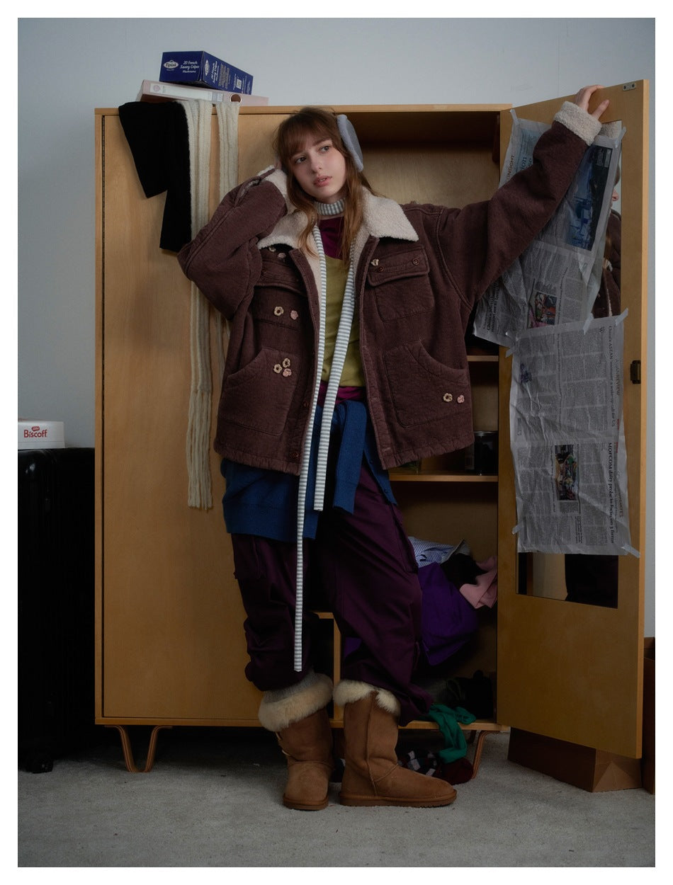 Model wearing a brown jacket with fleece-like collar and flower details, arm raised against an open wooden cabinet door