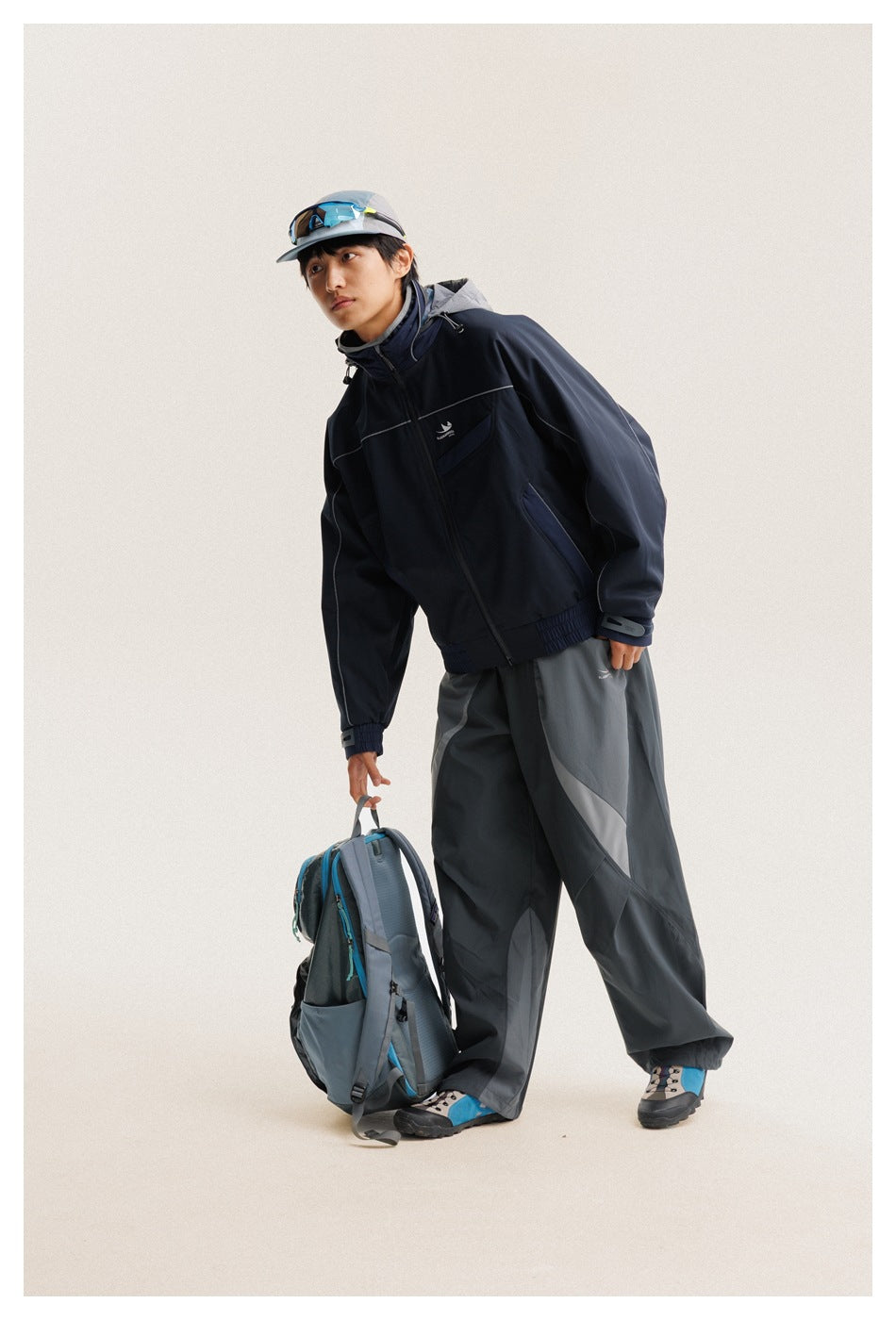 Model in a navy hooded jacket and gray paneled trousers, leaning beside a gray-blue backpack.
