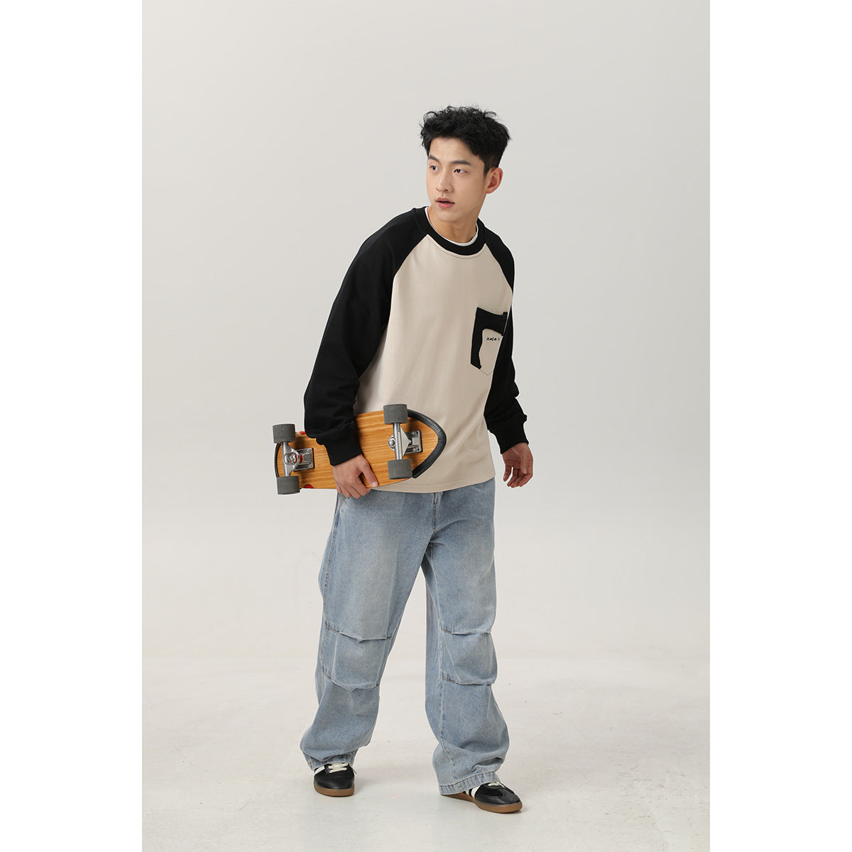 Model wearing a cream sweatshirt with black raglan sleeves and a chest pocket, paired with loose light-wash jeans.