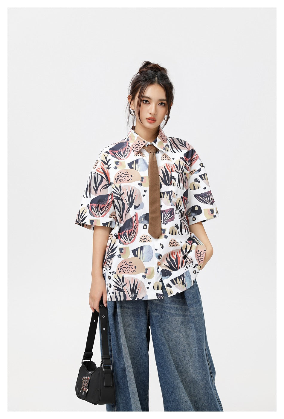 Closer front view of a model wearing a white botanical-print short-sleeve shirt with a brown tie and wide-leg jeans.