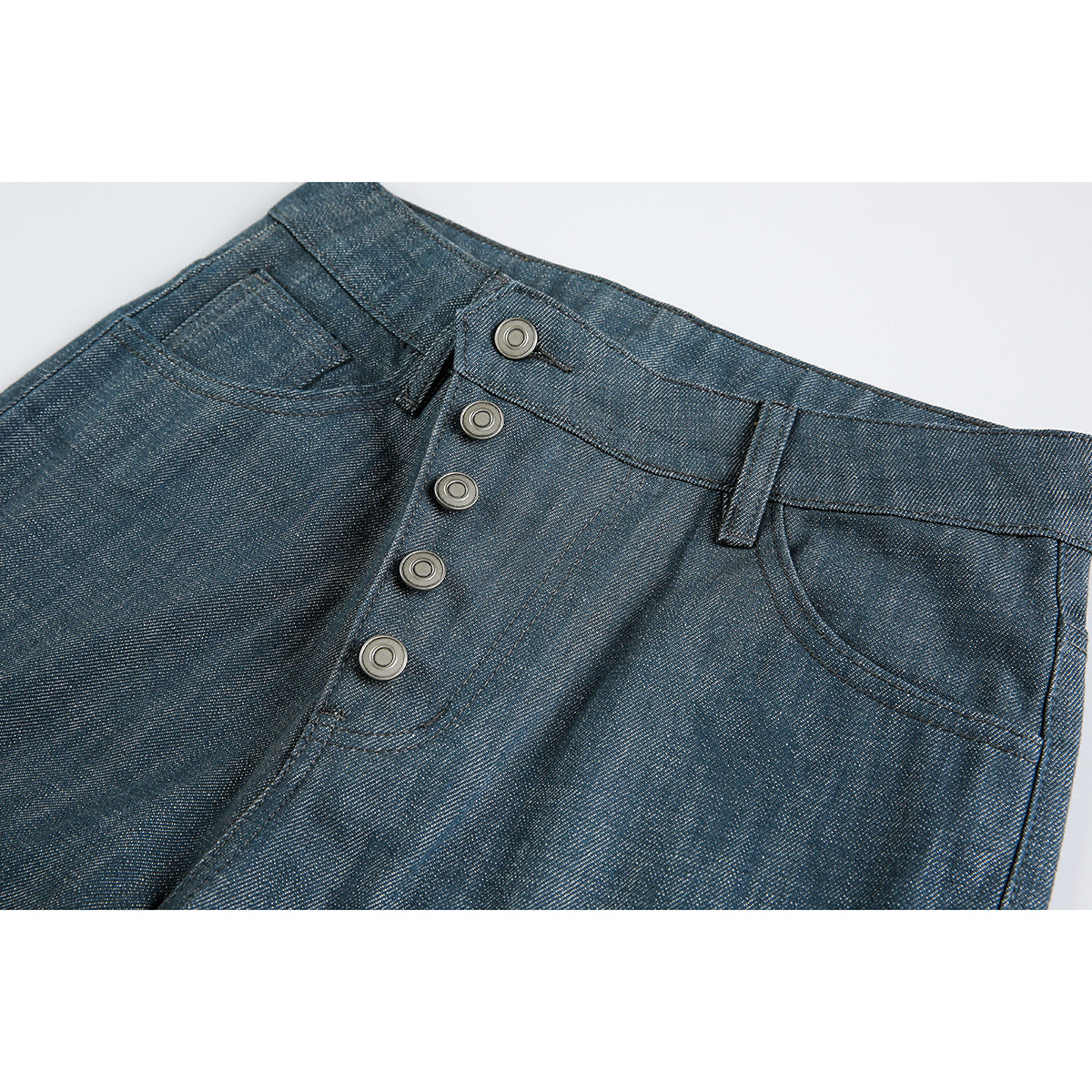 Close-up of blue-gray trousers showing the angled button fly, waistband, belt loop and front pocket.