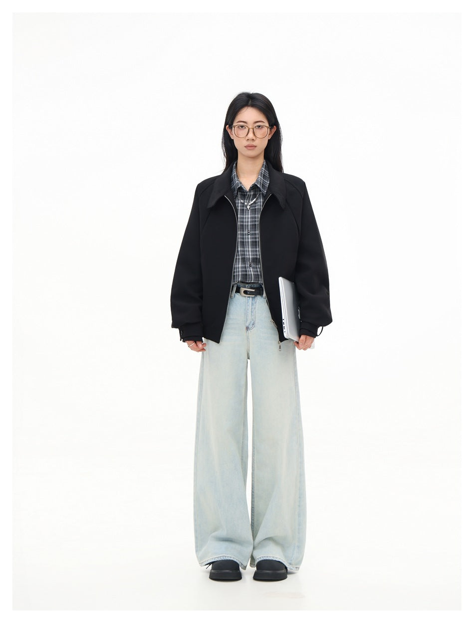 Model wearing black zip jacket over plaid shirt with light wide-leg jeans, front view, holding a laptop