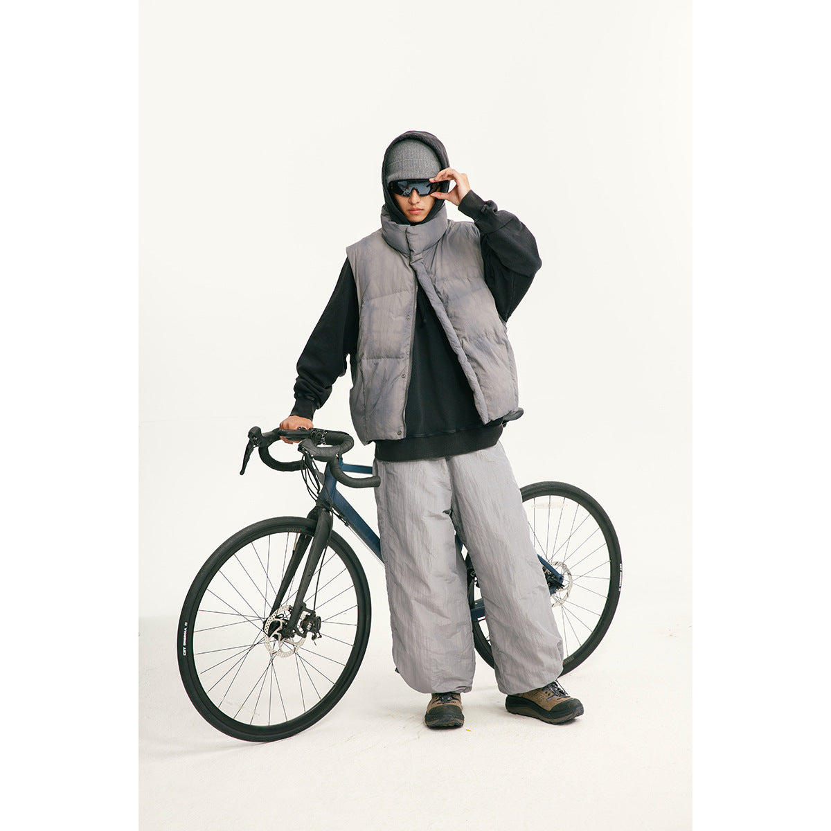 Model wearing a gray quilted vest with matching loose pants over a black hoodie, standing beside a bicycle.