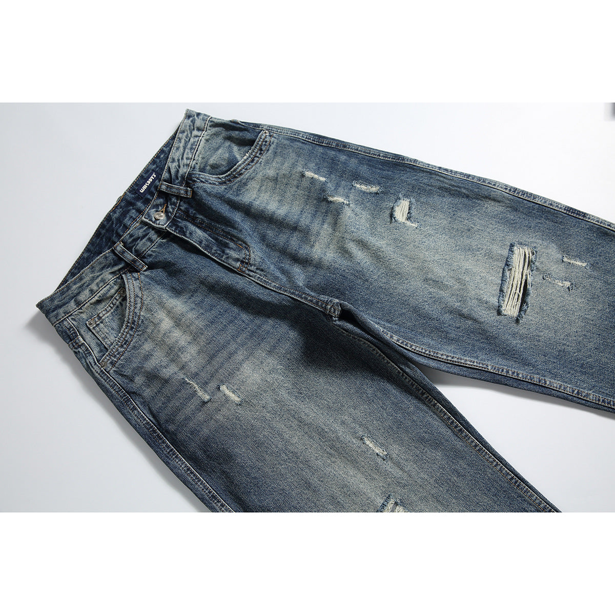 Front view of faded blue distressed jeans laid diagonally, with whiskering, ripped details, and five-pocket styling.