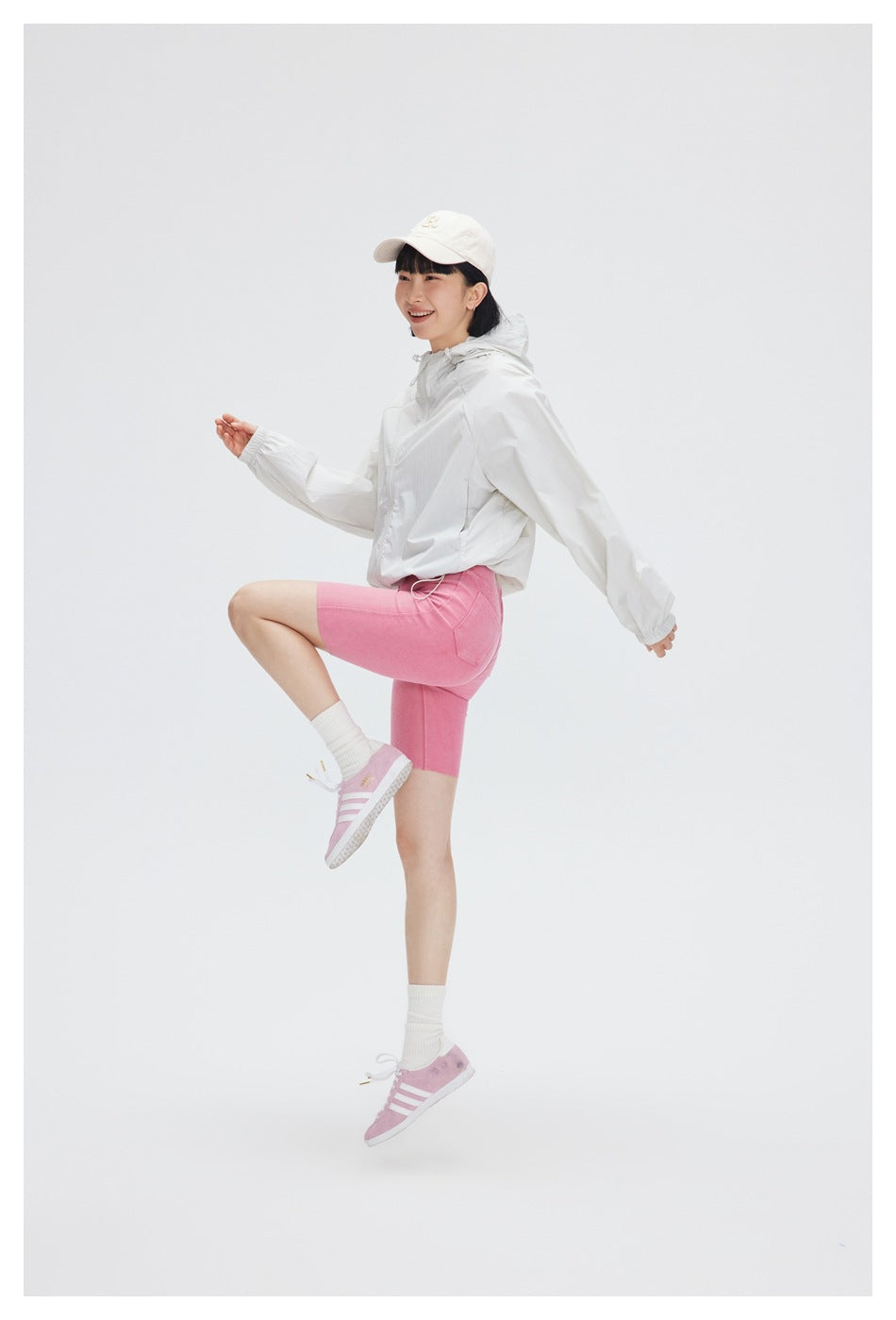 Model wearing a white hooded zip jacket and pink cycling shorts, posing with one knee raised.