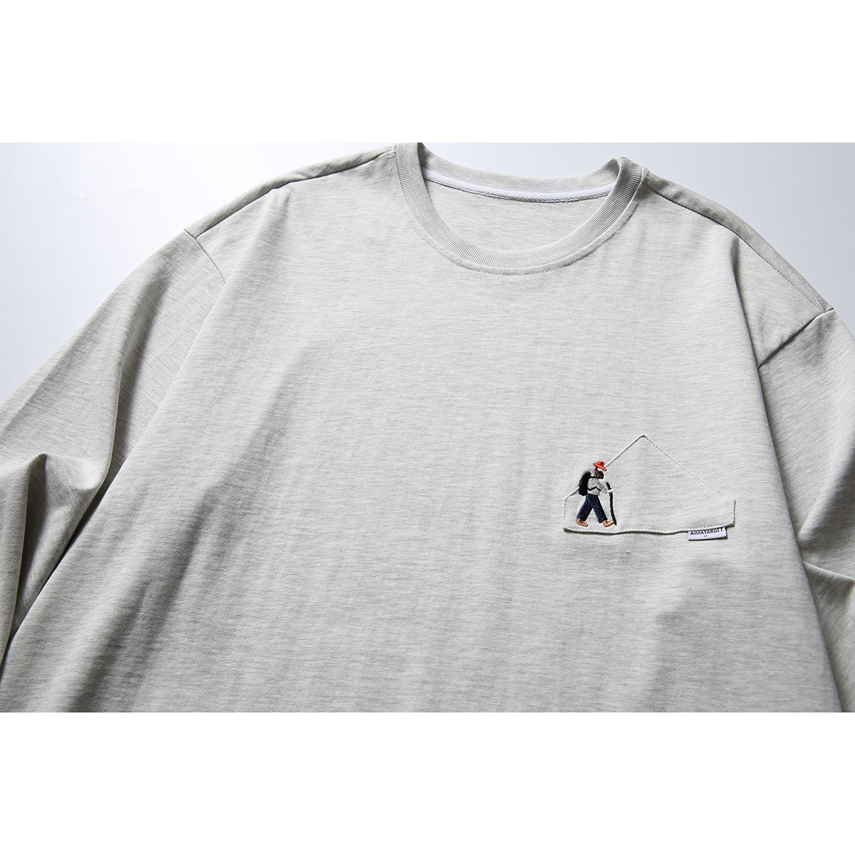 Close view of a light gray long-sleeve shirt with a small illustrated figure embroidered on a chest pocket.