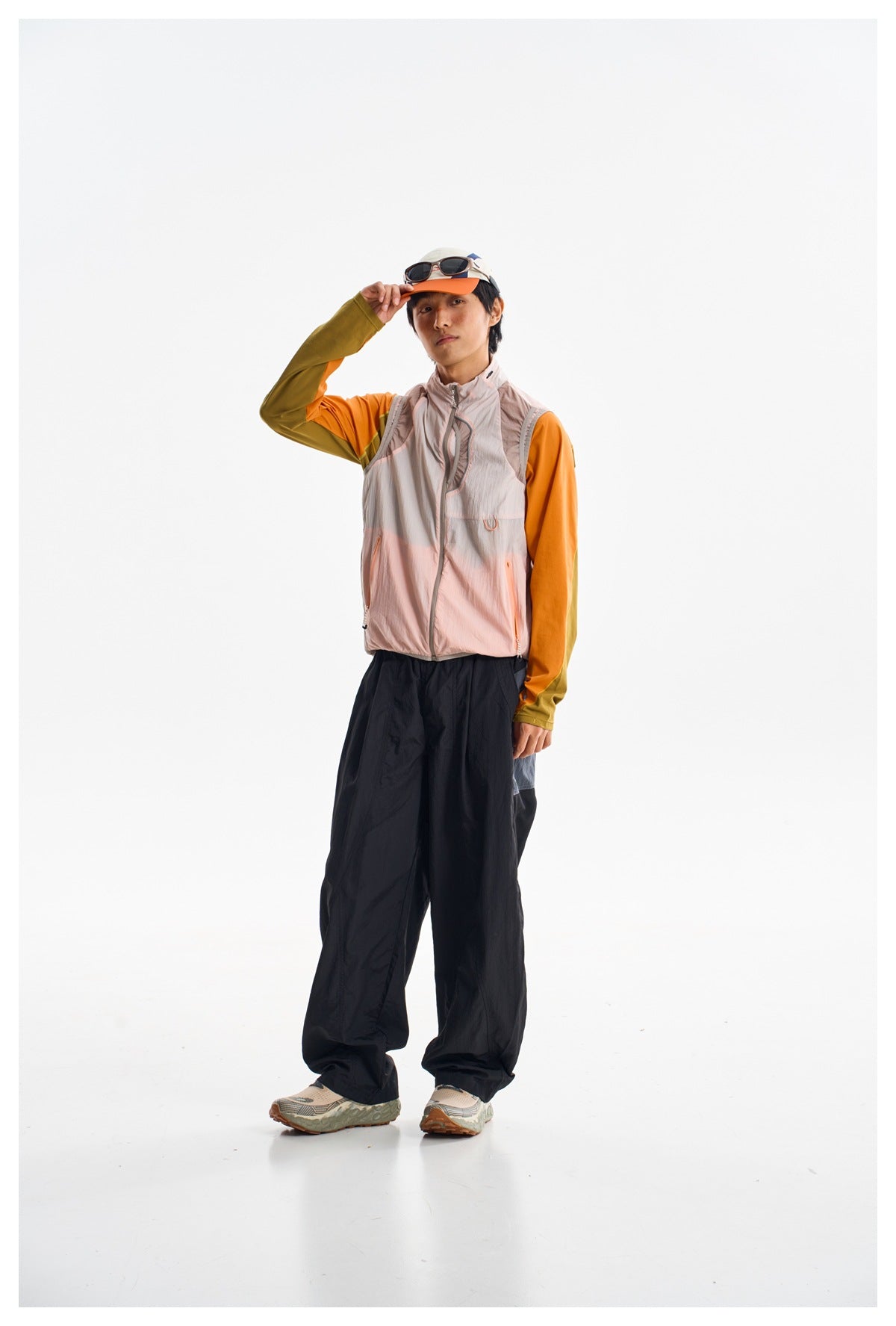Model wearing a pale pink zip-front vest over an orange and olive long-sleeve top with loose black pants.