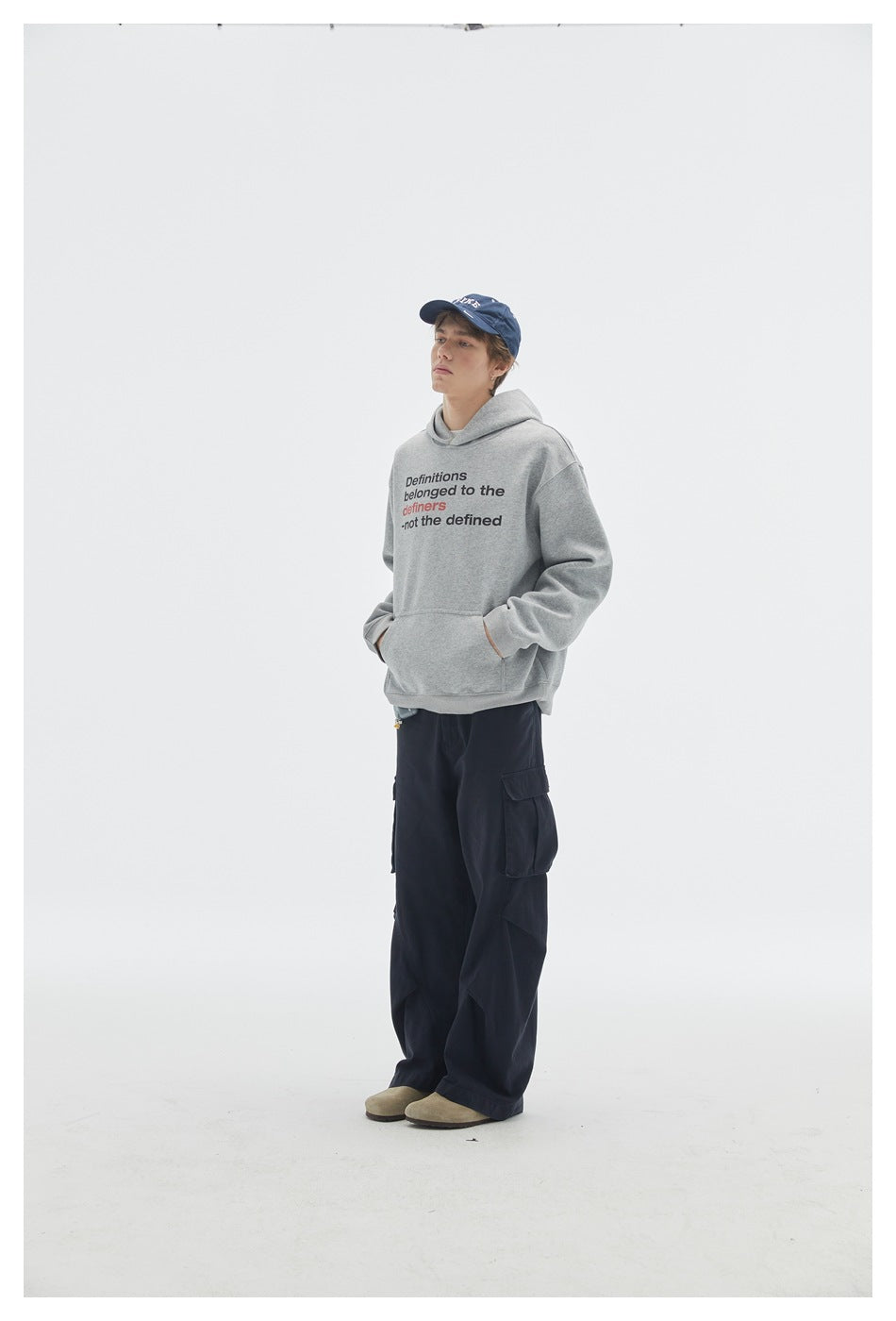 Angled full-length view of a model wearing a gray printed hoodie, navy cargo pants, and blue cap.