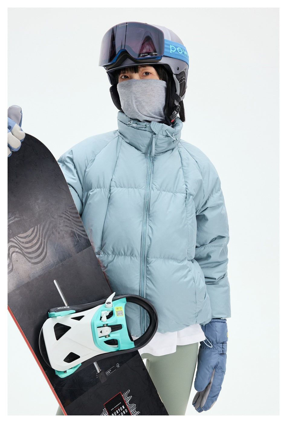 Person wearing a pale blue puffer jacket and ski gear, posed beside a snowboard with turquoise bindings.
