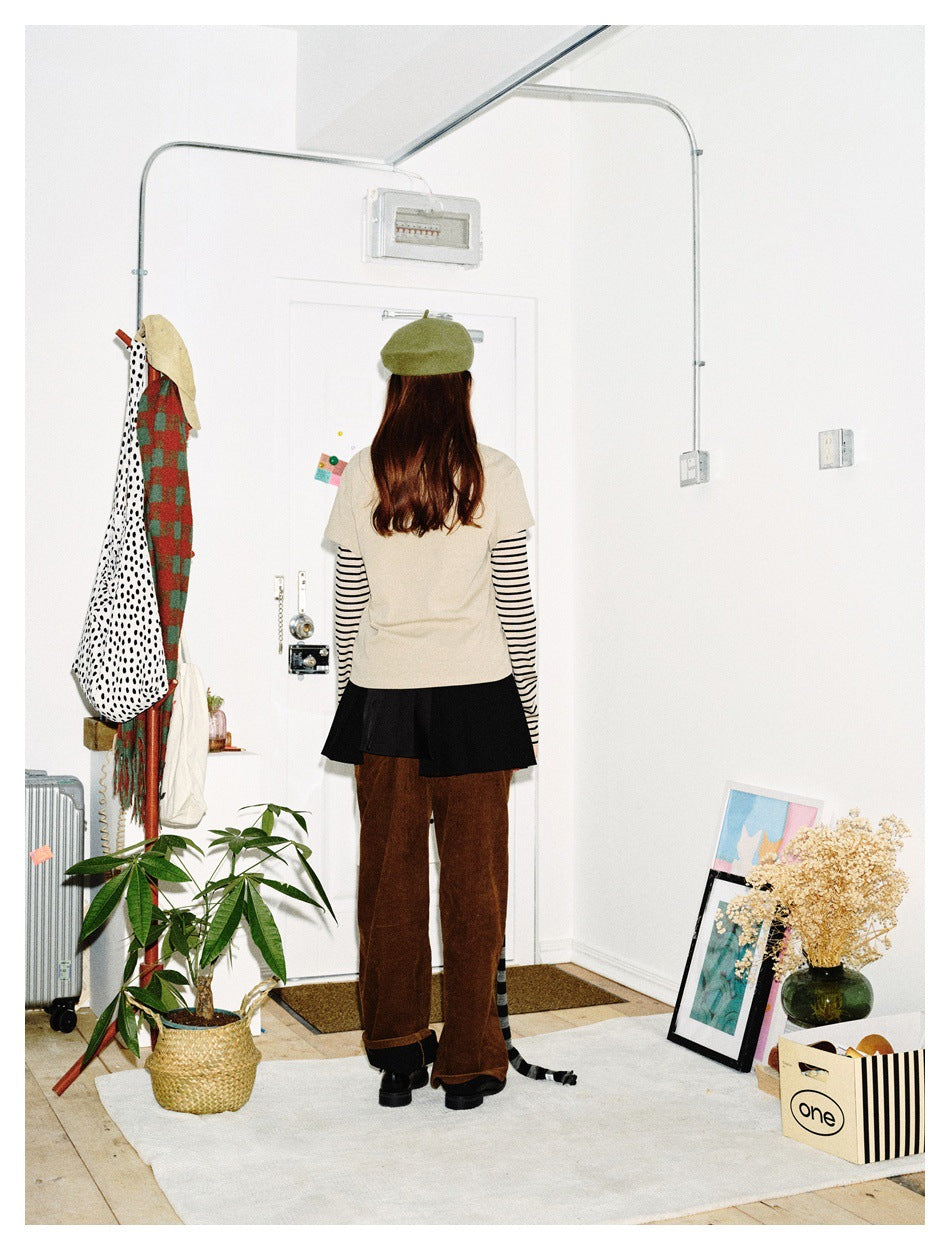 Back view of a model wearing a beige top over striped sleeves, black skirt, brown trousers and green beret; striped scarf