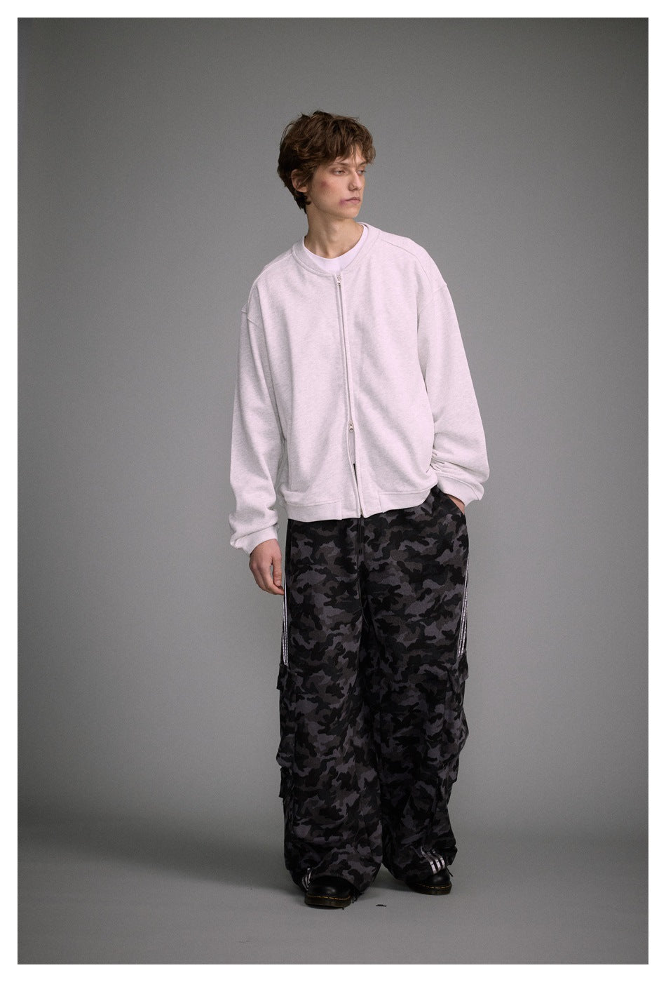 Full-length model wearing a light gray zip-front sweatshirt with loose dark camouflage cargo trousers.