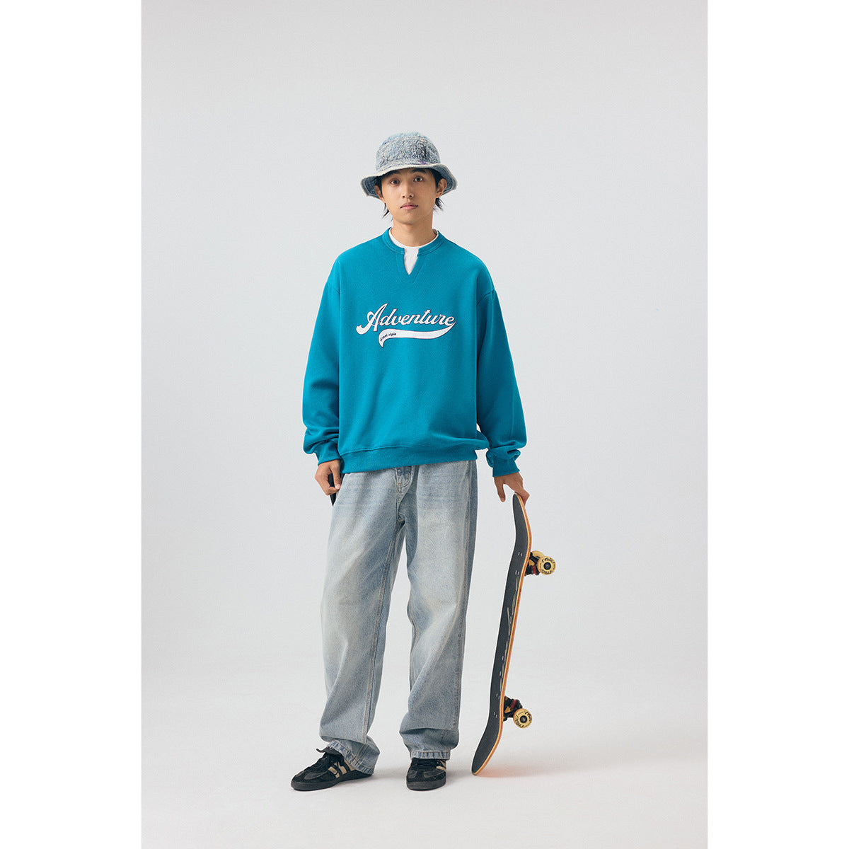 Model wearing a teal sweatshirt with a white script chest graphic, light jeans and a bucket hat.