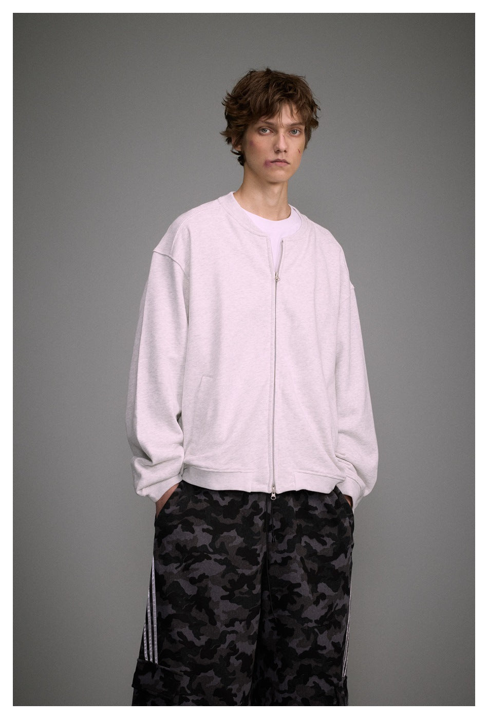 Closer view of a model wearing a light gray zip-front sweatshirt with loose dark camouflage trousers.