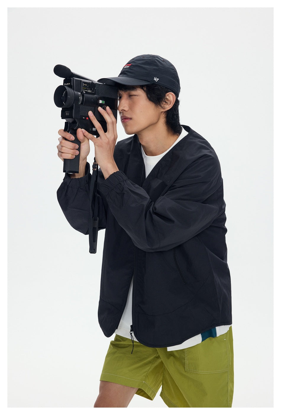 Model wearing a loose black zip-front jacket over a white shirt and lime-green shorts, holding a video camera.