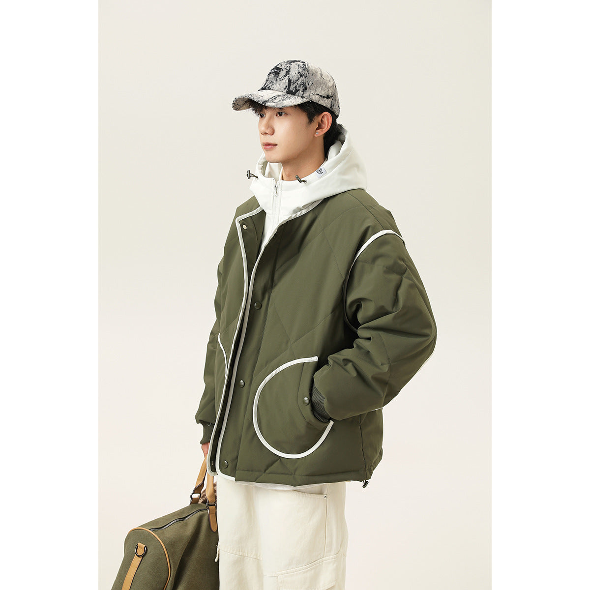 Model wearing an oversized olive hooded jacket with white piping, snap closures, and a large curved pocket.