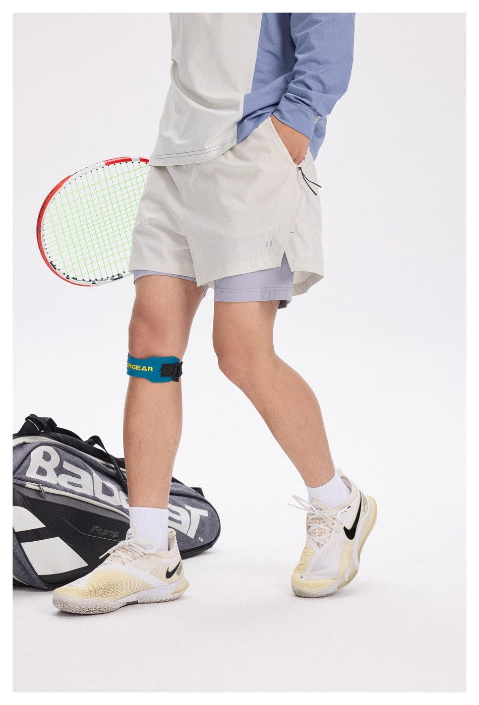 Front view of a model wearing light gray tennis shorts over lavender compression shorts, with a tennis racket beside them.