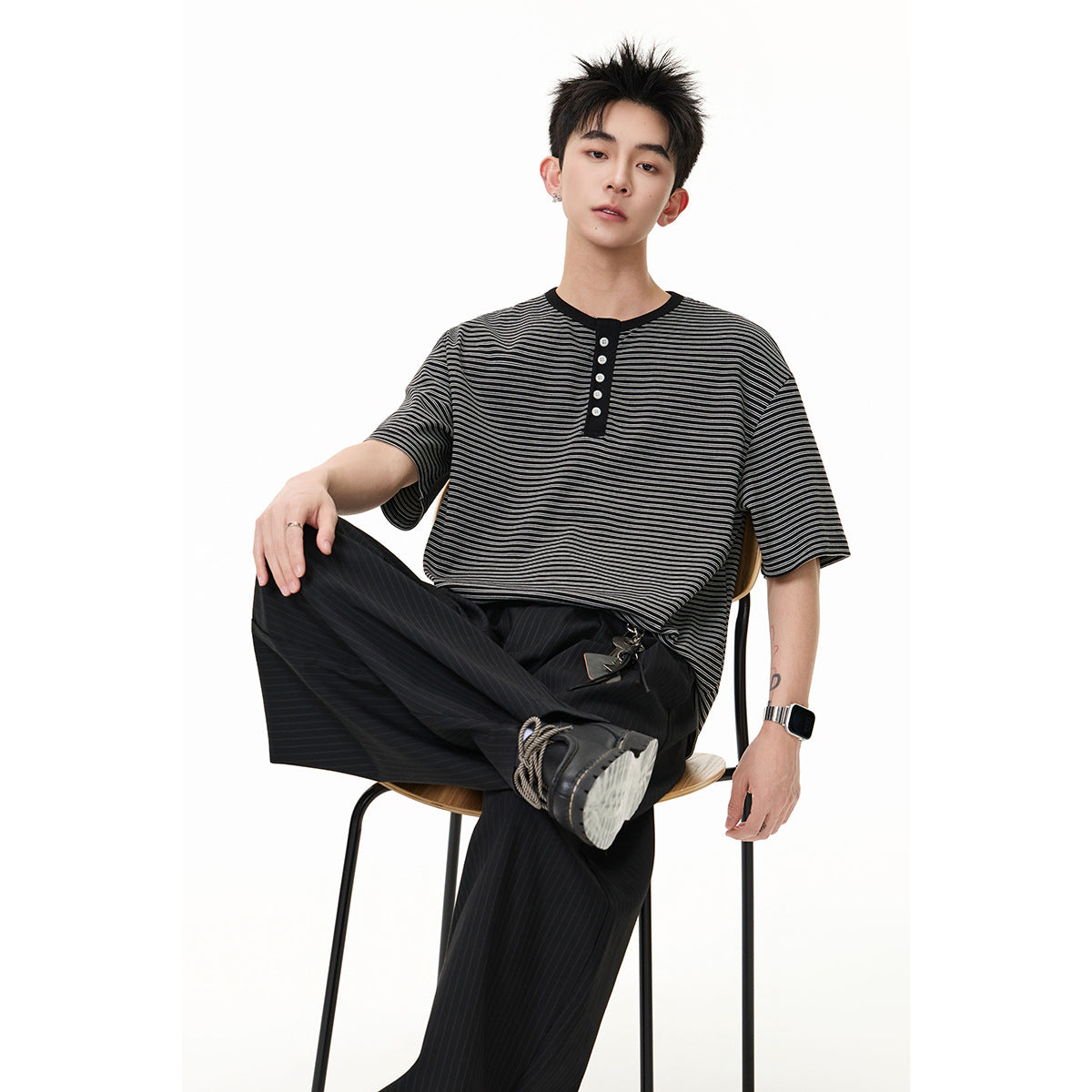 Seated model wearing a black and gray striped short-sleeve henley with black trousers.