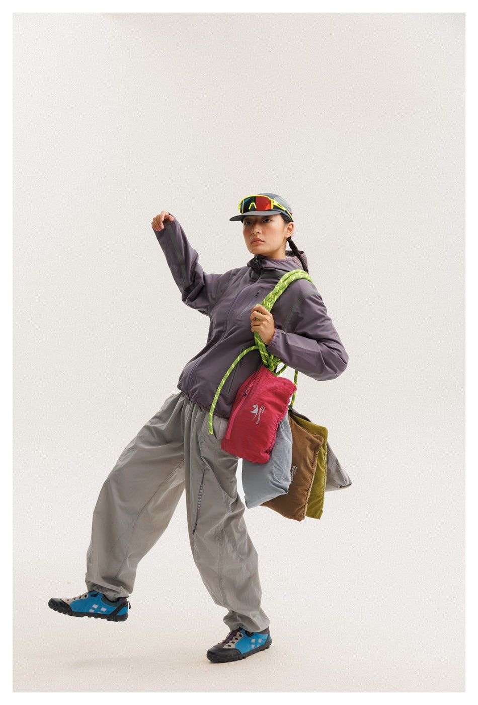 Model in a dark plum hooded jacket and gray pants, holding a bright green rope beside several colored bags.