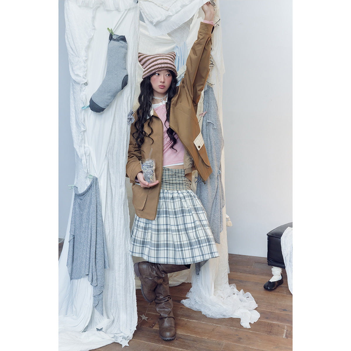 Model wears a brown jacket over a pink top and blue-gray plaid skirt, with a striped knit hat and brown boots.