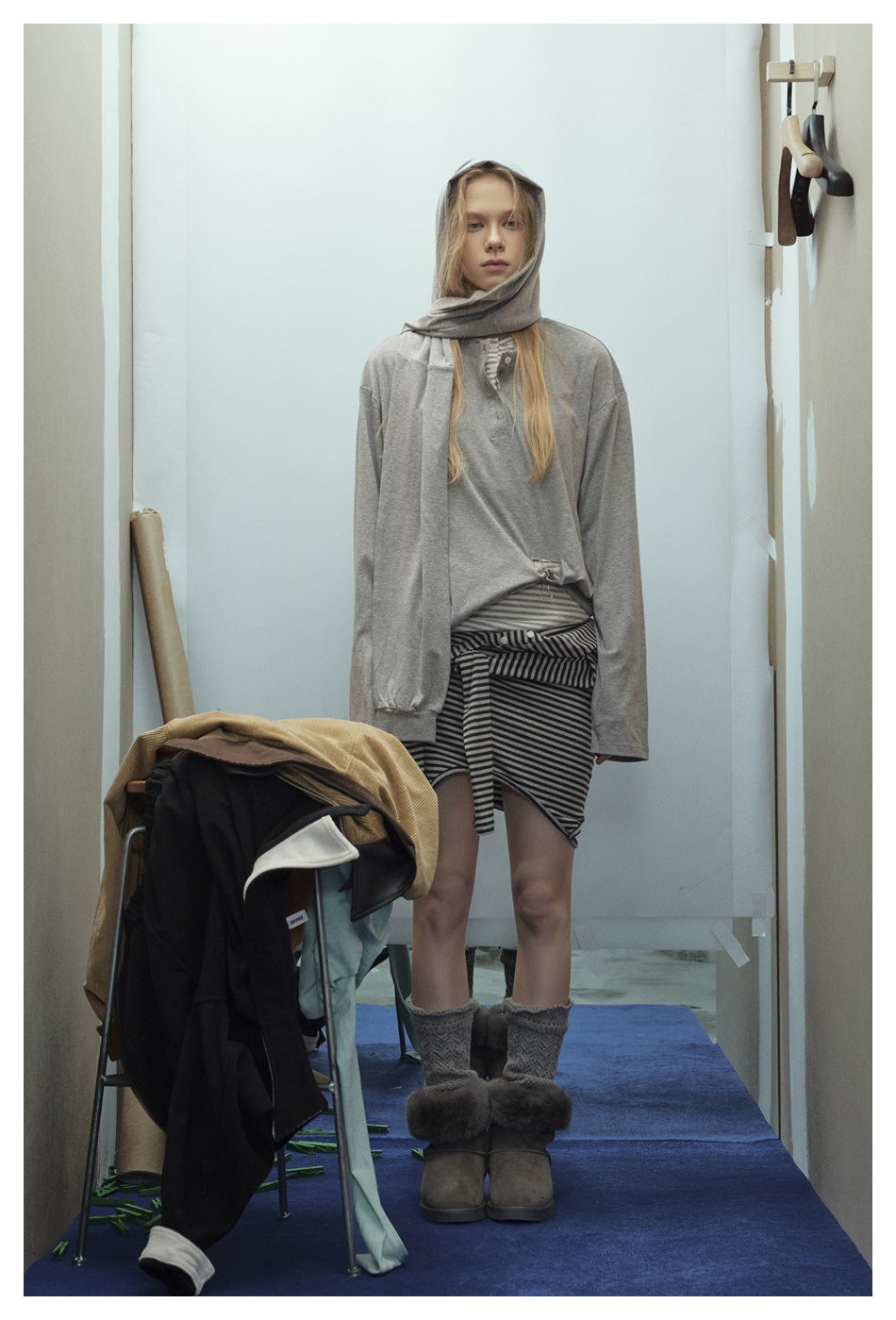 Model wearing a gray long-sleeve henley top with a shoulder layer draped over the head, styled with a striped layer and boots