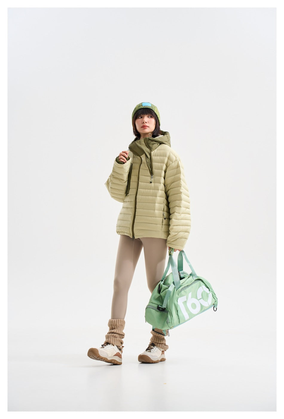 Full-length front view of a model wearing a cream quilted hooded jacket and carrying a green duffel bag.