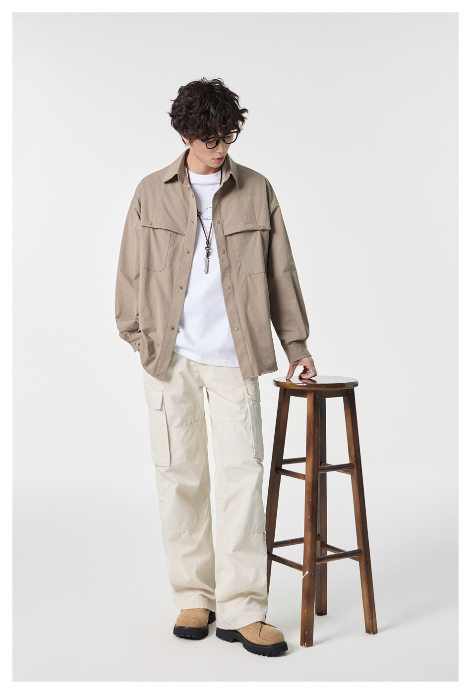 Full-length model wearing a taupe overshirt, white tee, and cream cargo pants beside a wooden stool.