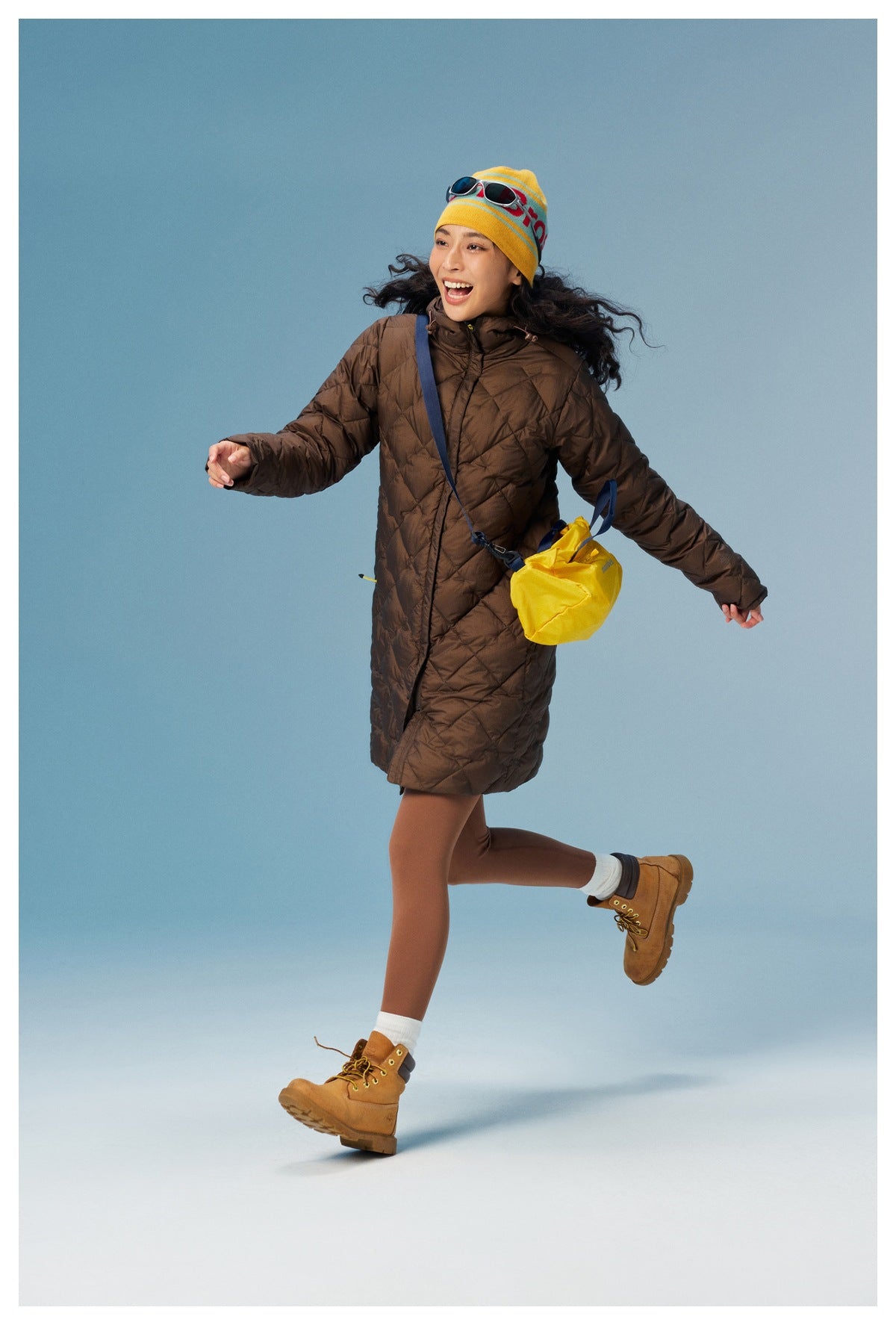 Model in a zipped brown quilted hooded coat, shown walking with a yellow crossbody bag.