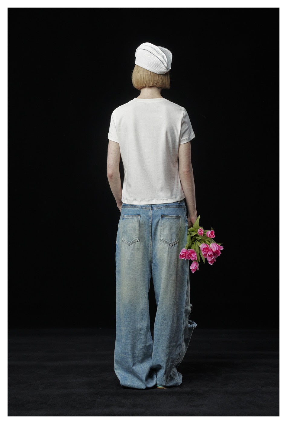 Back view of a model wearing a plain white T-shirt and loose blue jeans, holding pink tulips against black.