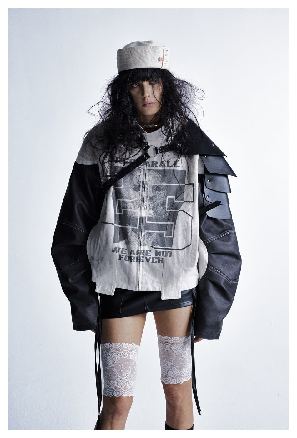 Model wearing the off-white and black jacket with a black skirt, lace leg wraps, hat and black shoulder armor accessory