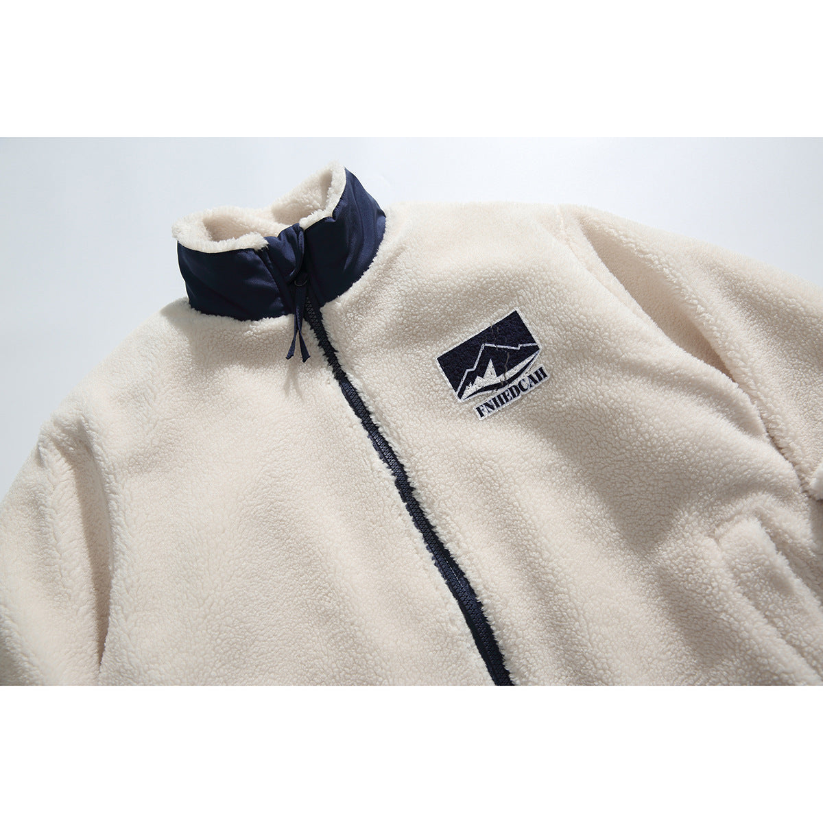 Close front view of an ivory pile fleece jacket with a navy stand collar, zipper, and chest patch.