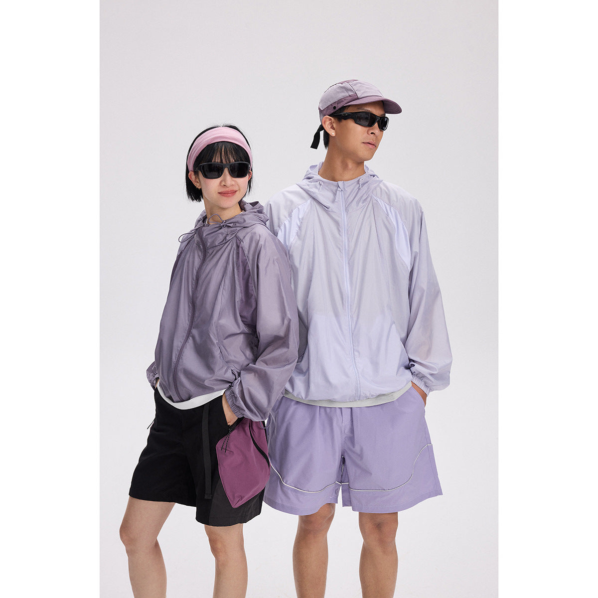 Two models wearing hooded zip jackets in charcoal and pale lavender, with matching shorts and sunglasses.