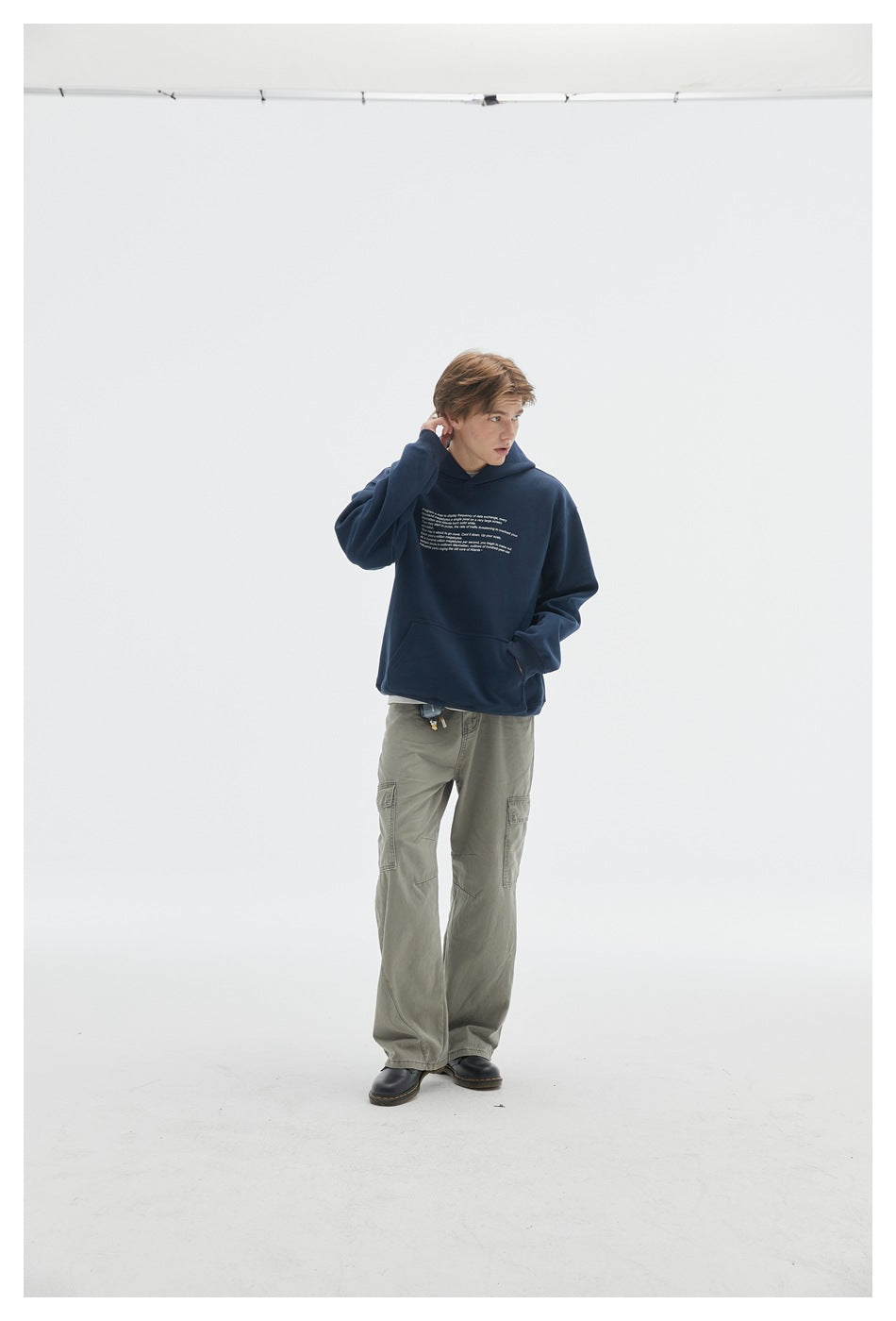 Model in a navy hoodie with small white chest text and olive cargo pants, posed with one hand near his neck.