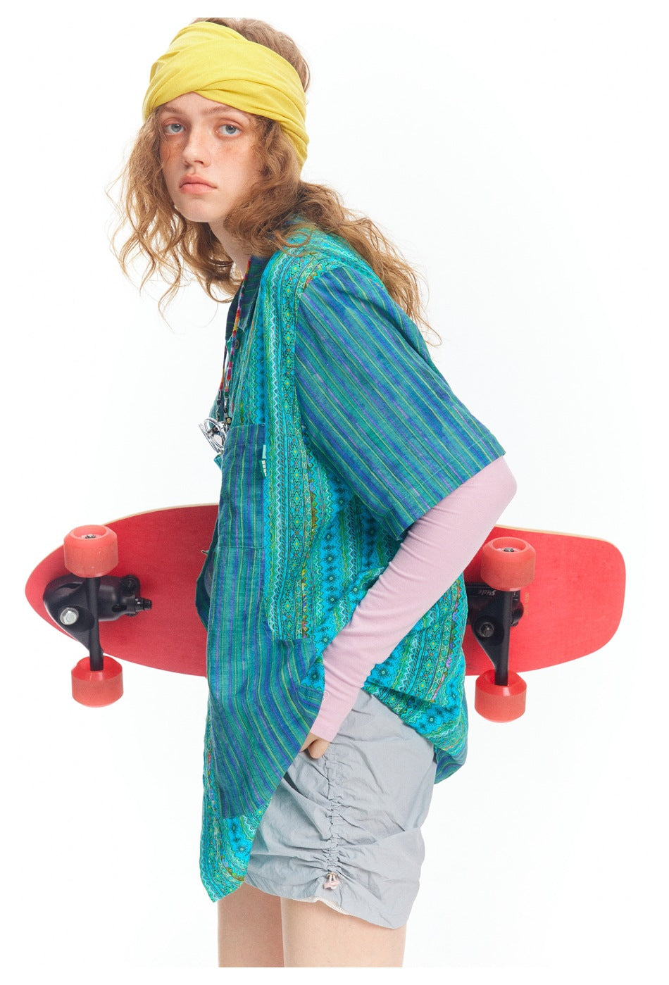 Side view of a model wearing a blue-green patterned short-sleeve shirt and holding a red skateboard.