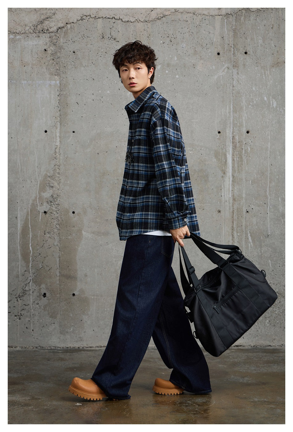 Model in a blue and black plaid shirt, shown in profile carrying a large black bag.
