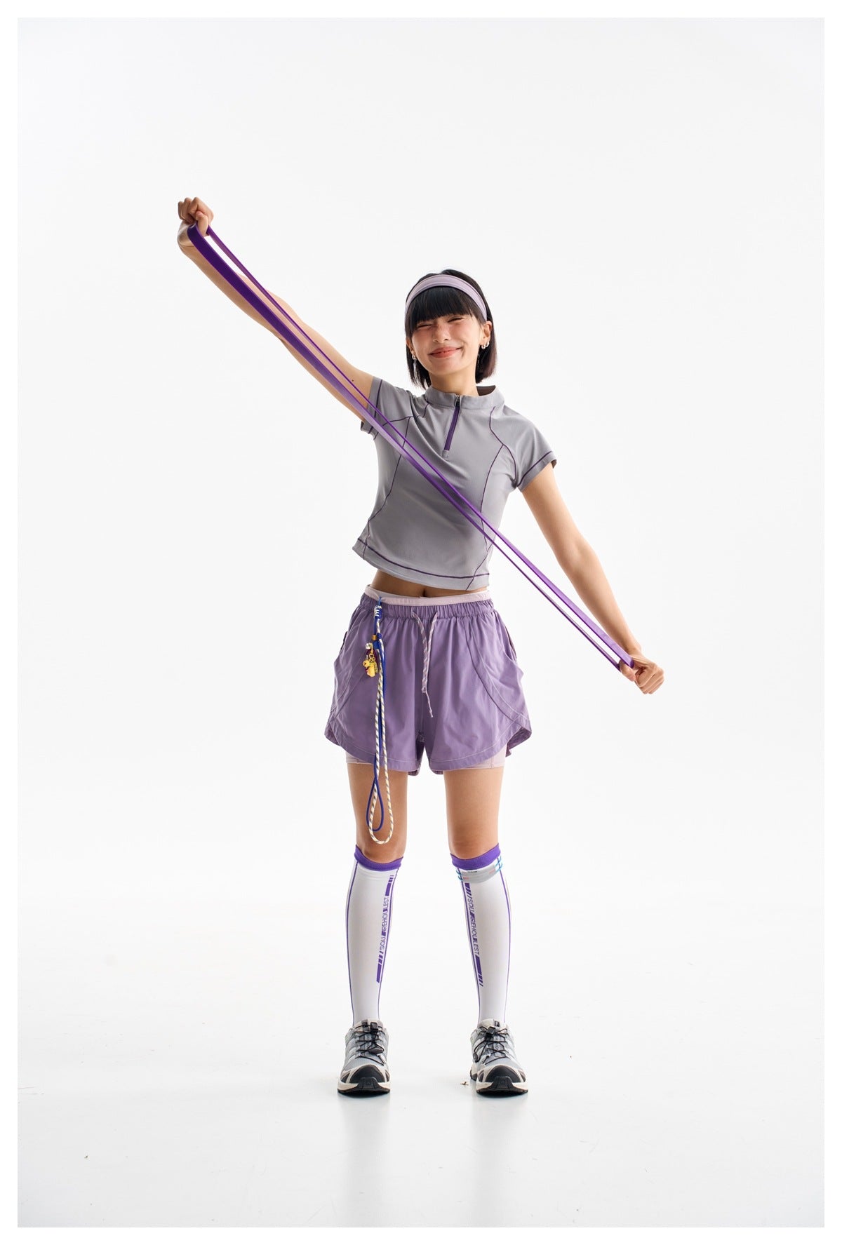 Model wearing a gray-lilac athletic top and purple shorts, stretching a purple resistance band diagonally across her body.