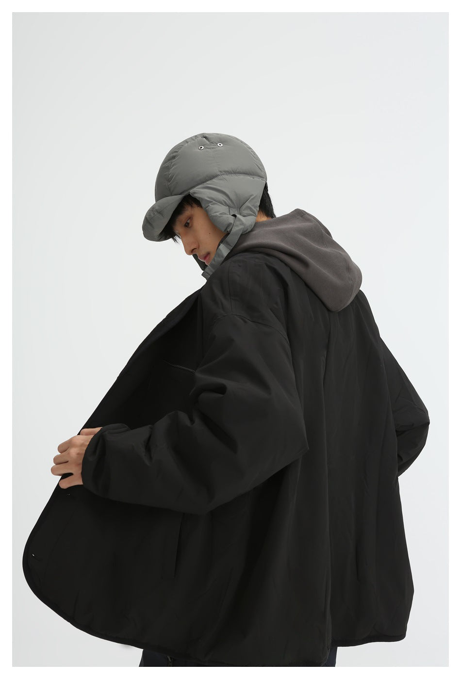 Rear three-quarter view of a model wearing a black oversized jacket, dark hoodie, and gray padded earflap cap.