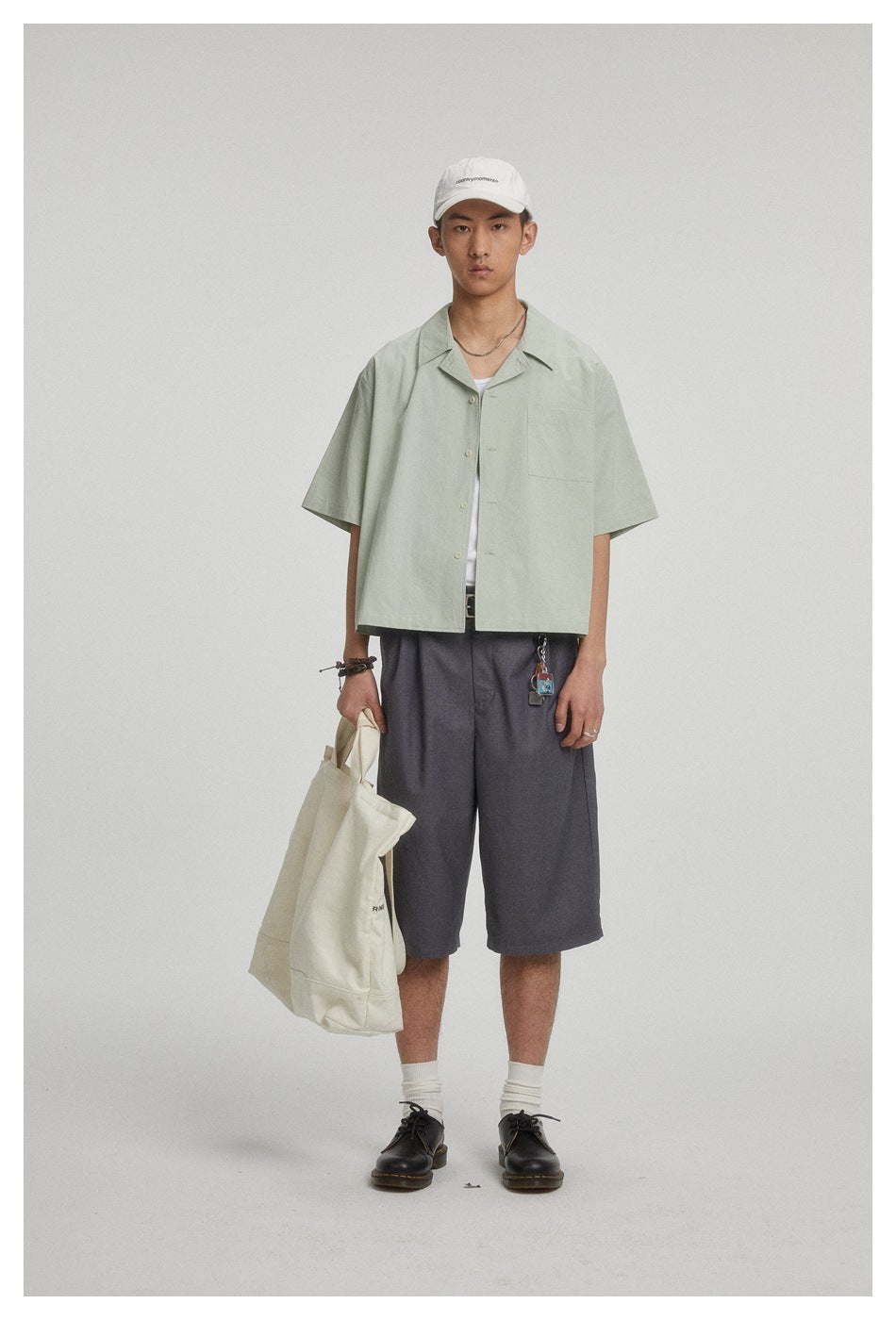 Model wearing a pale sage short-sleeve button-up shirt with a chest pocket and charcoal shorts.