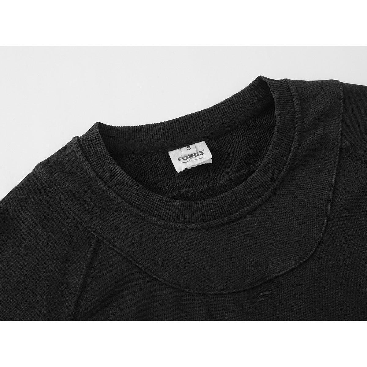 Close-up of a black crewneck sweatshirt showing its ribbed collar, curved chest seam, and small embroidered emblem.