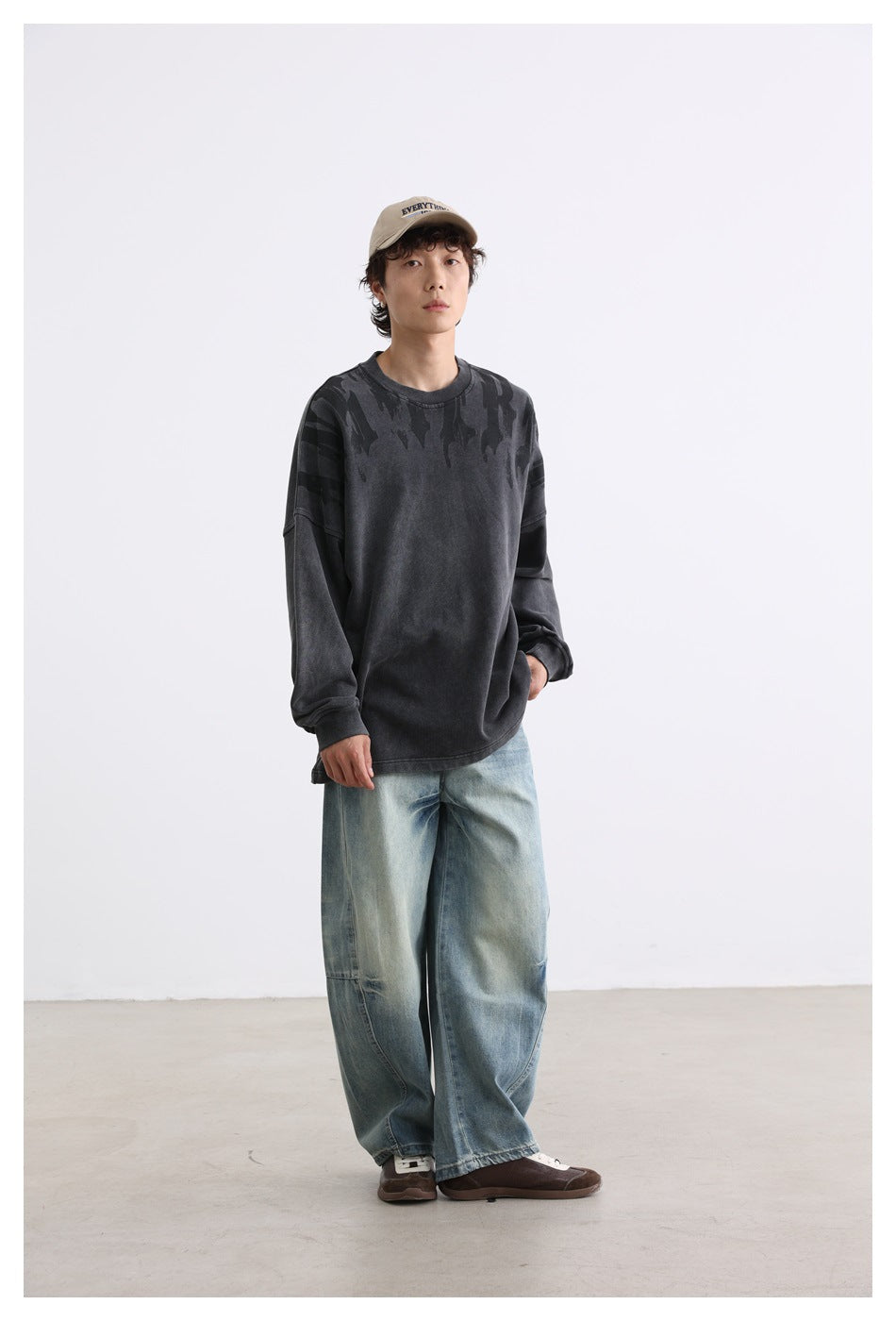 Full-length model wearing an oversized charcoal graphic-shoulder sweatshirt, loose light-wash jeans and a beige cap.