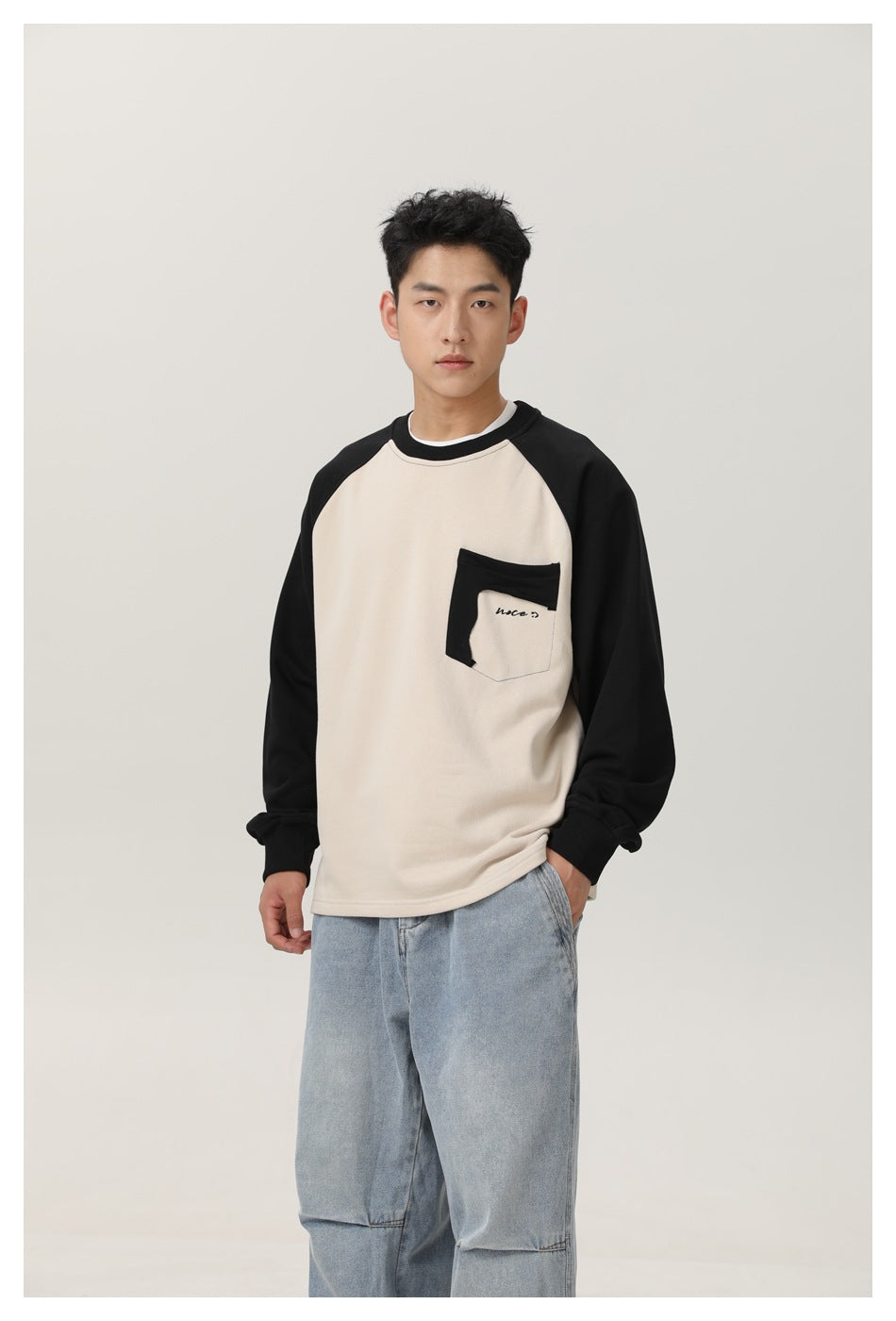 Front view of a model wearing a cream sweatshirt with black raglan sleeves and a black-trimmed chest pocket.
