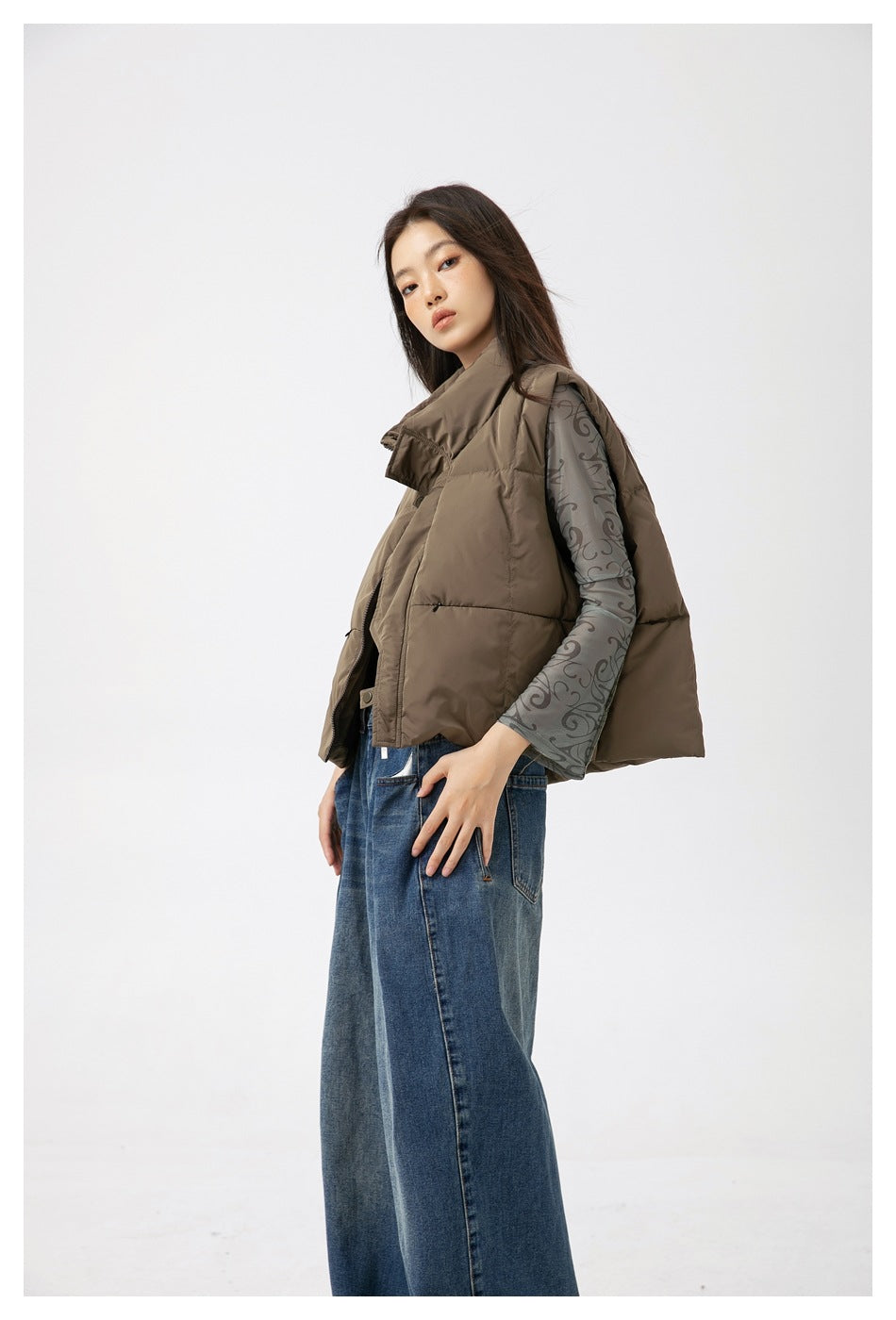 Woman in a gray-green quilted vest over a patterned mesh top and wide-leg jeans, shown from a three-quarter side angle