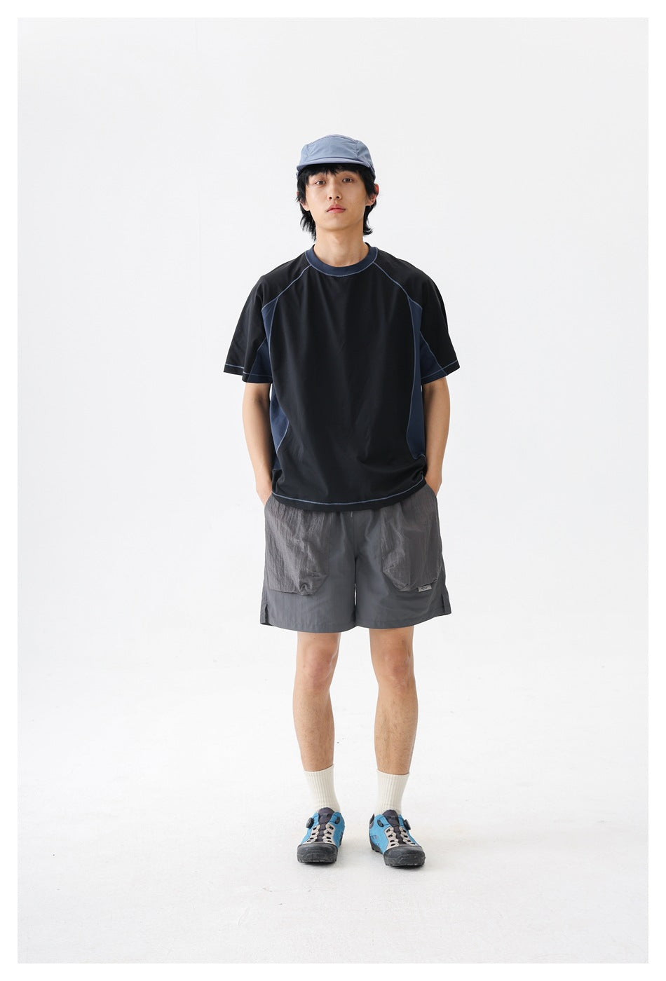 Front view of a man wearing a black T-shirt with navy panels, gray utility shorts and a blue-gray cap against a white