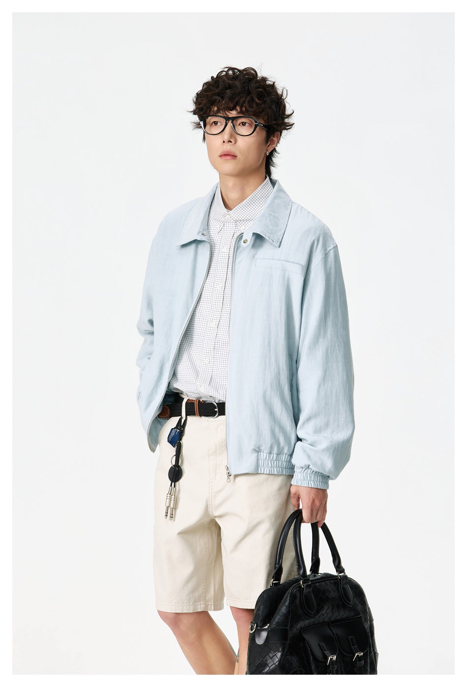 Model wearing a pale blue zip-front jacket with elasticated cuffs and hem, shown with a checked shirt and cream shorts.