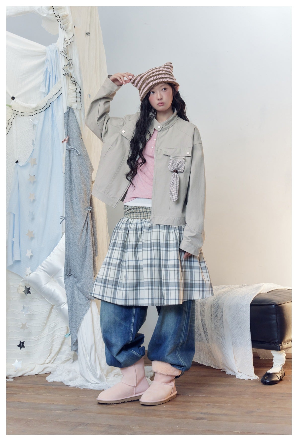 Model wears a light gray jacket, pink top, plaid skirt over jeans, and pale pink boots.