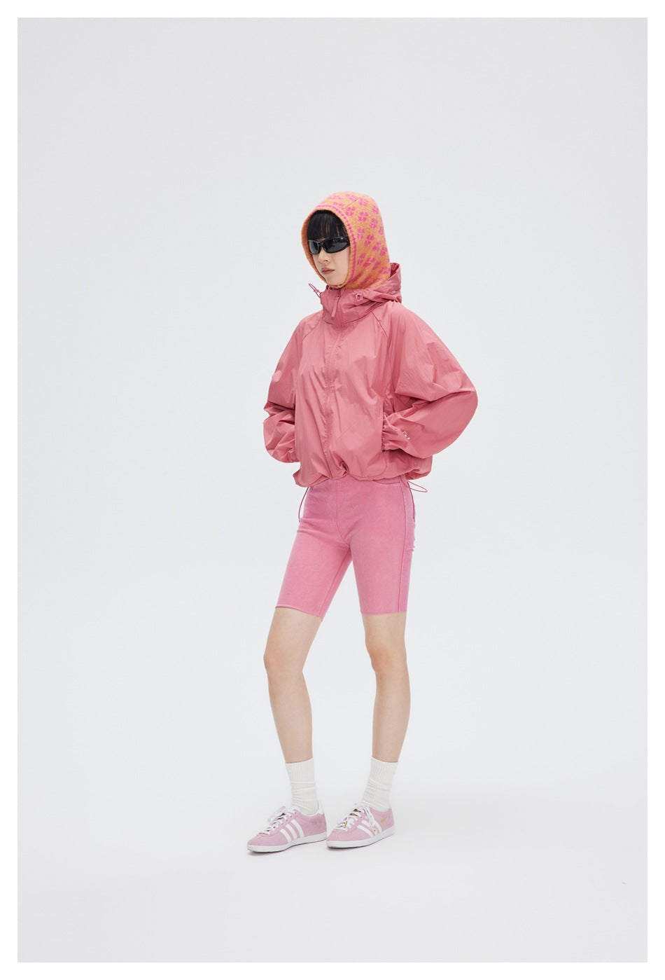 Person wearing a pink hooded zip jacket with a patterned head covering, sunglasses, pink shorts and sneakers